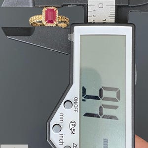 May include: A gold ring featuring a rectangular red gemstone surrounded by small white stones, held within the jaws of a black digital caliper. The digital display shows a measurement of 9.4 mm. The ring band has a textured, beaded surface design.