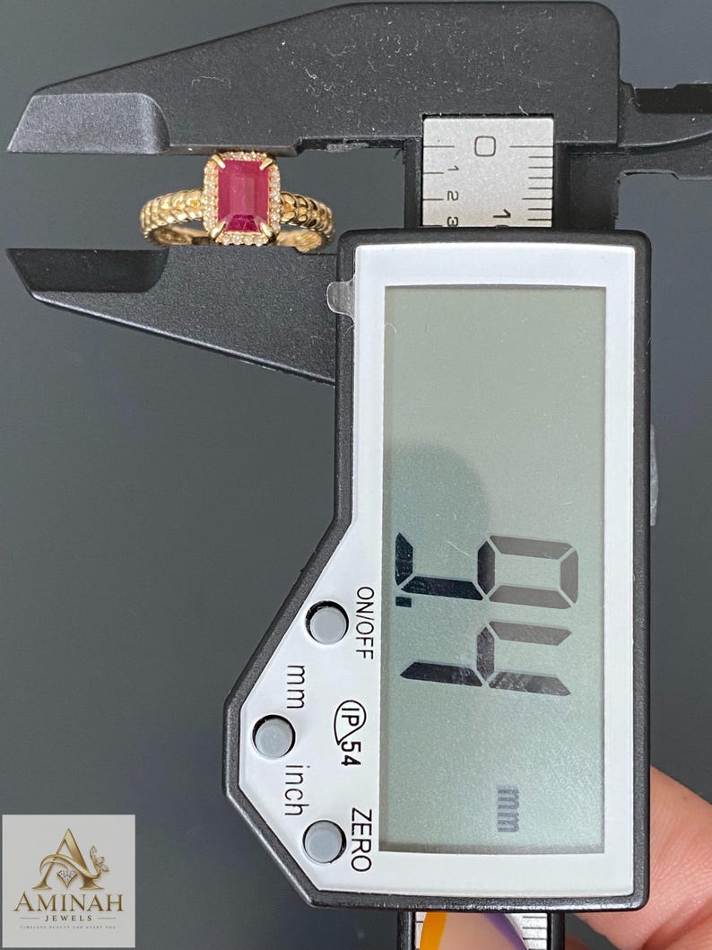May include: A gold ring featuring a rectangular red gemstone surrounded by small white stones, held within the jaws of a black digital caliper. The digital display shows a measurement of 9.4 mm. The ring band has a textured, beaded surface design.