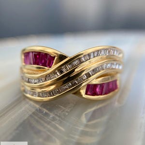 May include: A gold ring featuring a crossover design with rows of channel-set baguette-cut rubies and clear baguette-cut diamonds. The band has a polished finish and the logo for Aminah Jewels is visible in the bottom left corner of the image.