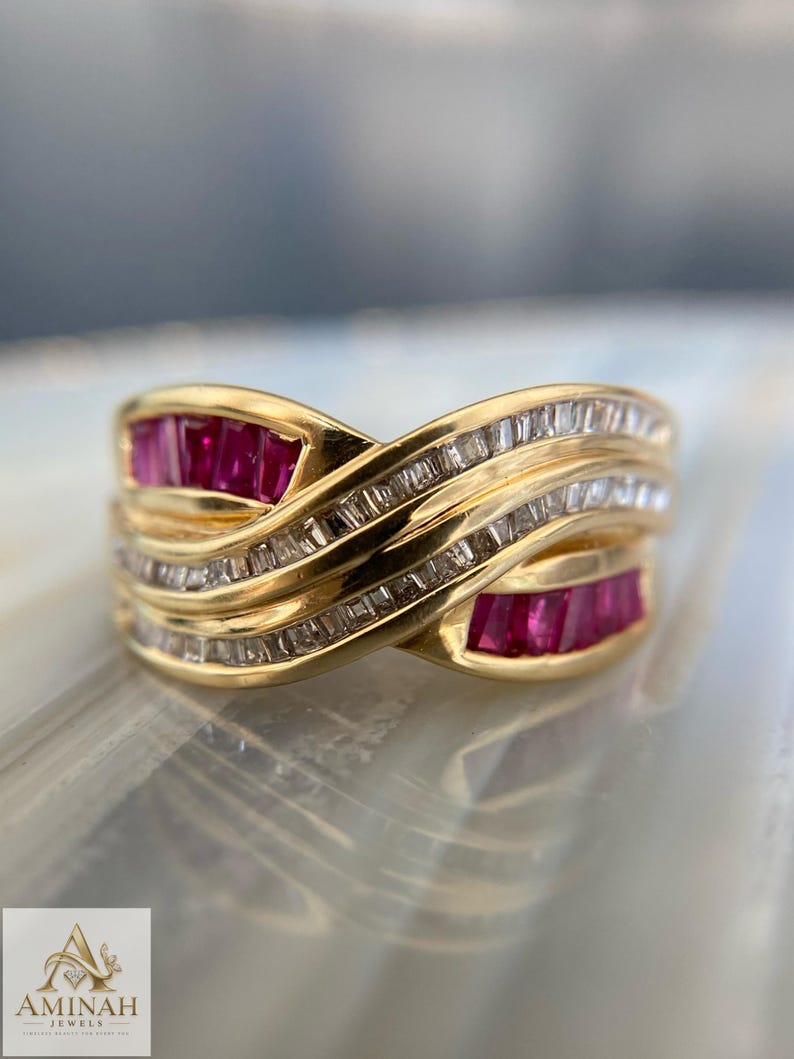 K&ouml;nnte beinhalten: Ein Goldring mit einem &uuml;berkreuzten Design, das Reihen von im Kanal gefassten Baguette-Rubinen und klaren Baguette-Diamanten aufweist. Das Band hat eine polierte Oberfl&auml;che und das Logo von Aminah Jewels ist unten links im Bild zu sehen.