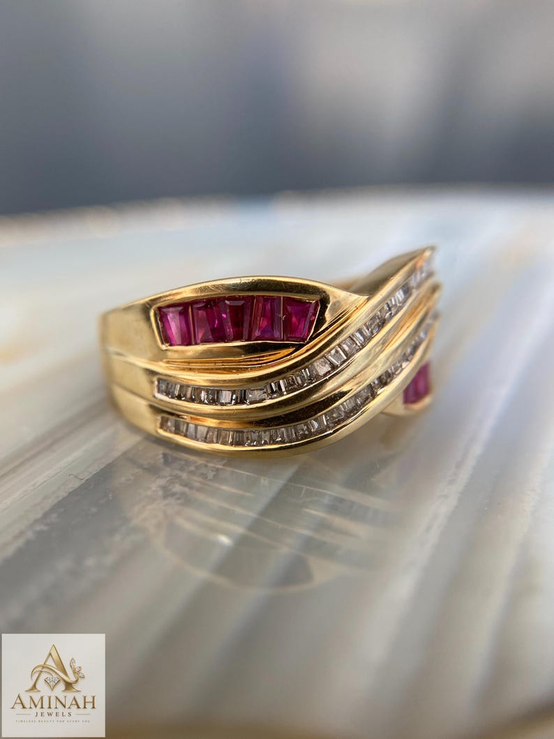K&ouml;nnte beinhalten: Ein Goldring mit einer geschwungenen Reihe aus f&uuml;nf rechteckigen roten Rubinen und zwei parallelen Reihen kleiner, im Kanal gefasster, klarer Baguette-Diamanten. Das Band hat eine polierte Goldoberfl&auml;che mit einem wellenf&ouml;rmigen Design.