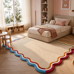 May include: A rectangular area rug with a solid beige center and a scalloped, wavy border featuring layered bands of orange, white, and blue colors. The rug has a soft, plush texture and is positioned on a light-colored wooden floor.