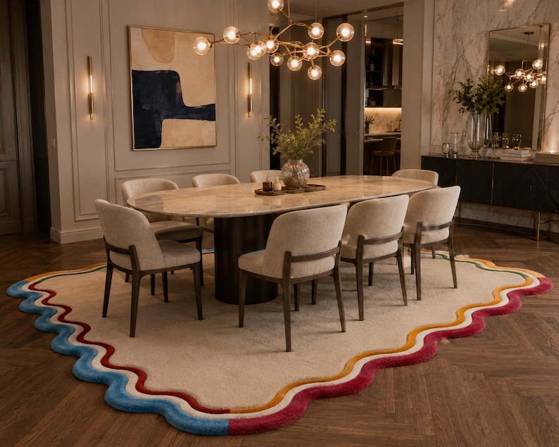 May include: A large, irregularly shaped area rug featuring a beige center and a wavy, multi-colored border in shades of blue, yellow, orange, and magenta. The rug is positioned beneath an oval dining table with six upholstered chairs.