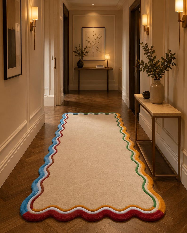 May include: A rectangular runner rug with a beige center and a scalloped edge featuring concentric bands of blue, white, green, yellow, and red. The rug is positioned on a dark wood herringbone floor in a hallway with cream-colored walls.