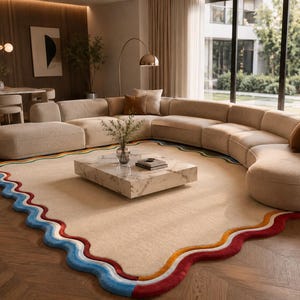 May include: A large, rectangular area rug with a cream-colored center and a wavy, multi-colored border featuring thick bands of blue, red, and orange. The rug is positioned on a wooden floor, partially underneath a large, curved, beige modular sofa.