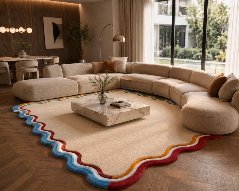 May include: A large, rectangular area rug with a cream-colored center and a wavy, multi-colored border featuring thick bands of blue, red, and orange. The rug is positioned on a wooden floor, partially underneath a large, curved, beige modular sofa.