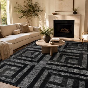 Könnte beinhalten: Ein modernes Wohnzimmer mit einem großen, geometrisch gemusterten Teppich in Schwarz- und Grautönen. Der Teppich ist der Mittelpunkt, ergänzt durch ein beiges Sofa, einen Holztisch und einen Kamin. Der Raum ist hell und natürlich belichtet.