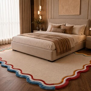 May include: A rectangular area rug with a cream-colored center and a scalloped border featuring layered bands of blue, white, orange, and red. The rug is positioned on a wooden floor beneath a bed with a beige upholstered frame and bedding.