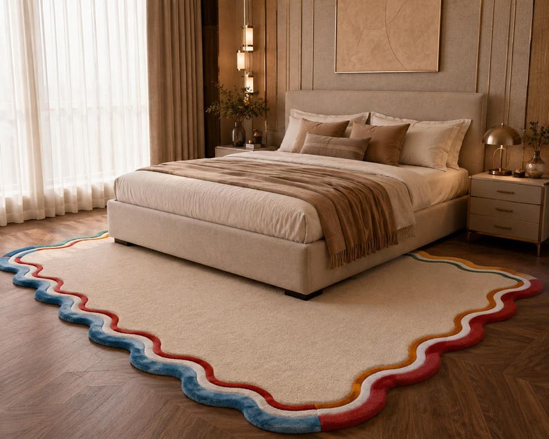 May include: A rectangular area rug with a cream-colored center and a scalloped border featuring layered bands of blue, white, orange, and red. The rug is positioned on a wooden floor beneath a bed with a beige upholstered frame and bedding.