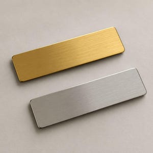 May include: Two rectangular name badges with rounded corners. One badge has a brushed gold finish, and the other has a brushed silver finish. Both items feature a flat, metallic surface with a horizontal grain texture and a clean, simple design.
