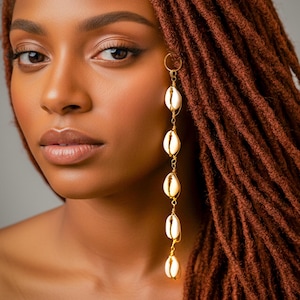 May include: A long, dangling earring featuring five white cowrie shells arranged vertically. Each shell is individually wrapped in thin, gold-toned wire and connected by small gold-toned metal links, ending in a circular hoop at the top for attachment.
