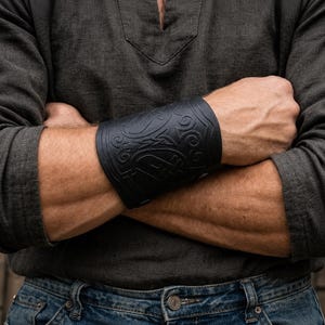 May include: Black leather wrist cuff with an embossed Celtic knot design. The cuff is worn on the forearm and features intricate swirling patterns and a central knot. The cuff is a dark, matte black and appears to be made of leather.
