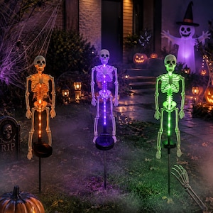 May include: Three standing skeleton garden stakes with illuminated spines. The left skeleton glows orange, the center skeleton glows purple, and the right skeleton glows green. Each figure is mounted on a black stake with a solar panel base.