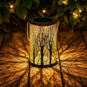 May include: A cylindrical solar lantern with a dark metal frame featuring a cut-out tree branch pattern. The lantern emits a warm, golden light from within, casting intricate, elongated tree-shaped shadows across the surface beneath it.