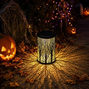 May include: A cylindrical solar lantern featuring a black metal cutout design of bare trees. The lantern emits a warm yellow light that projects a detailed, branch-like shadow pattern onto the ground, surrounded by fallen autumn leaves.