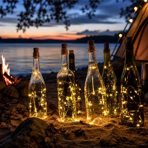 May include: Five clear glass bottles with cork stoppers, each containing a string of warm white fairy lights. The bottles are arranged in a row on a sandy surface, glowing brightly against a twilight background with a campfire and a tent.