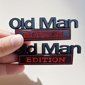 Pu&ograve; includere: Due emblemi neri "Old Man Edition". Il testo "Old Man" &egrave; in grassetto, lettere maiuscole sopra la parola "Edition" in rosso, il tutto su sfondo rettangolare nero.