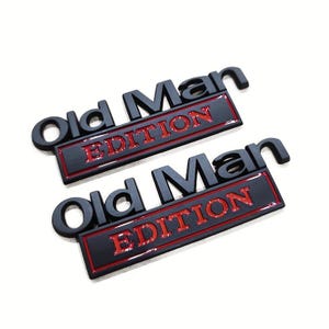 Pu&ograve; includere: Due emblemi neri "Old Man Edition" con scritte rosse. Il testo &egrave; in rilievo e ha un effetto 3D. Gli emblemi sono rettangolari con angoli arrotondati, su sfondo bianco.