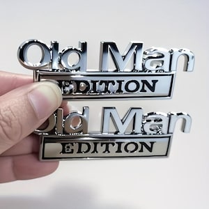 Pu&ograve; includere: Due emblemi metallici cromati con la scritta "Old Man EDITION" in grassetto, senza grazie. La parola "EDITION" &egrave; su uno sfondo rettangolare argentato. Gli emblemi sono tenuti da una mano.