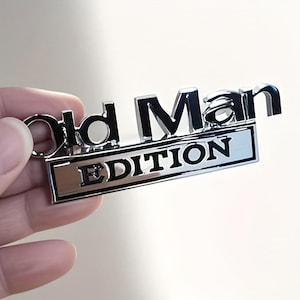 Pu&ograve; includere: Un portachiavi argentato lucido con le parole "Old Man EDITION" in lettere nere in rilievo. Il portachiavi ha una base rettangolare con la parola "EDITION" e un piccolo anello per le chiavi.