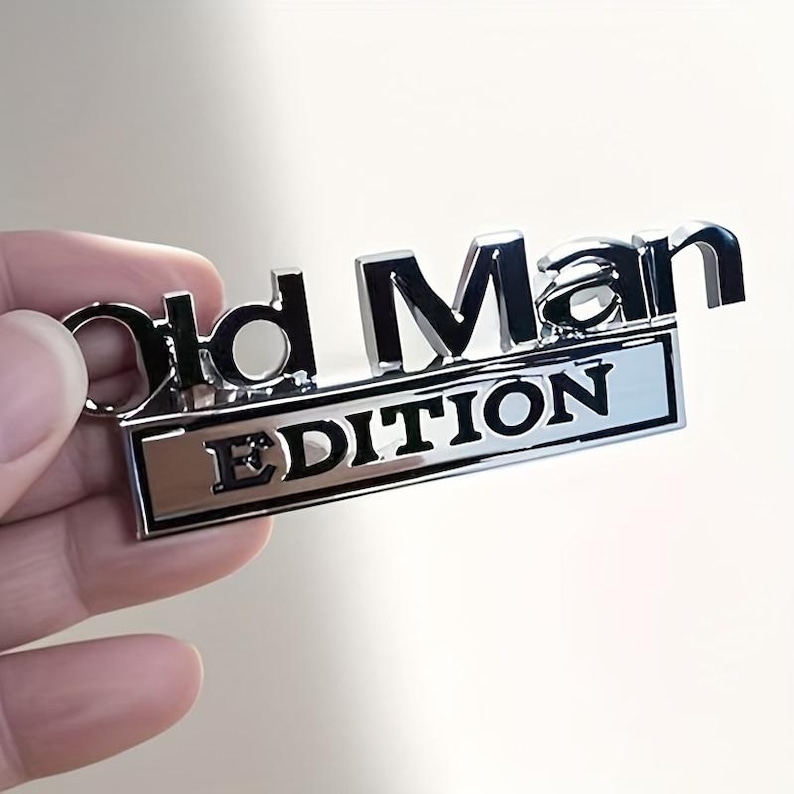 Pu&ograve; includere: Un portachiavi argentato lucido con le parole "Old Man EDITION" in lettere nere in rilievo. Il portachiavi ha una base rettangolare con la parola "EDITION" e un piccolo anello per le chiavi.