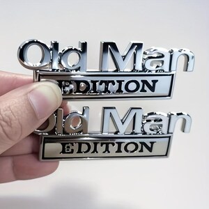 Pu&ograve; includere: Due emblemi metallici cromati con la scritta "Old Man EDITION". La parola "EDITION" &egrave; su uno sfondo rettangolare argentato. Gli emblemi sono tenuti in mano, su sfondo chiaro.