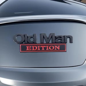 Pu&ograve; includere: Un'auto grigio scuro con un emblema nero che recita "Old Man EDITION" in grassetto. La parola "EDITION" &egrave; in un rettangolo rosso, aggiungendo un colore contrastante al design generale.
