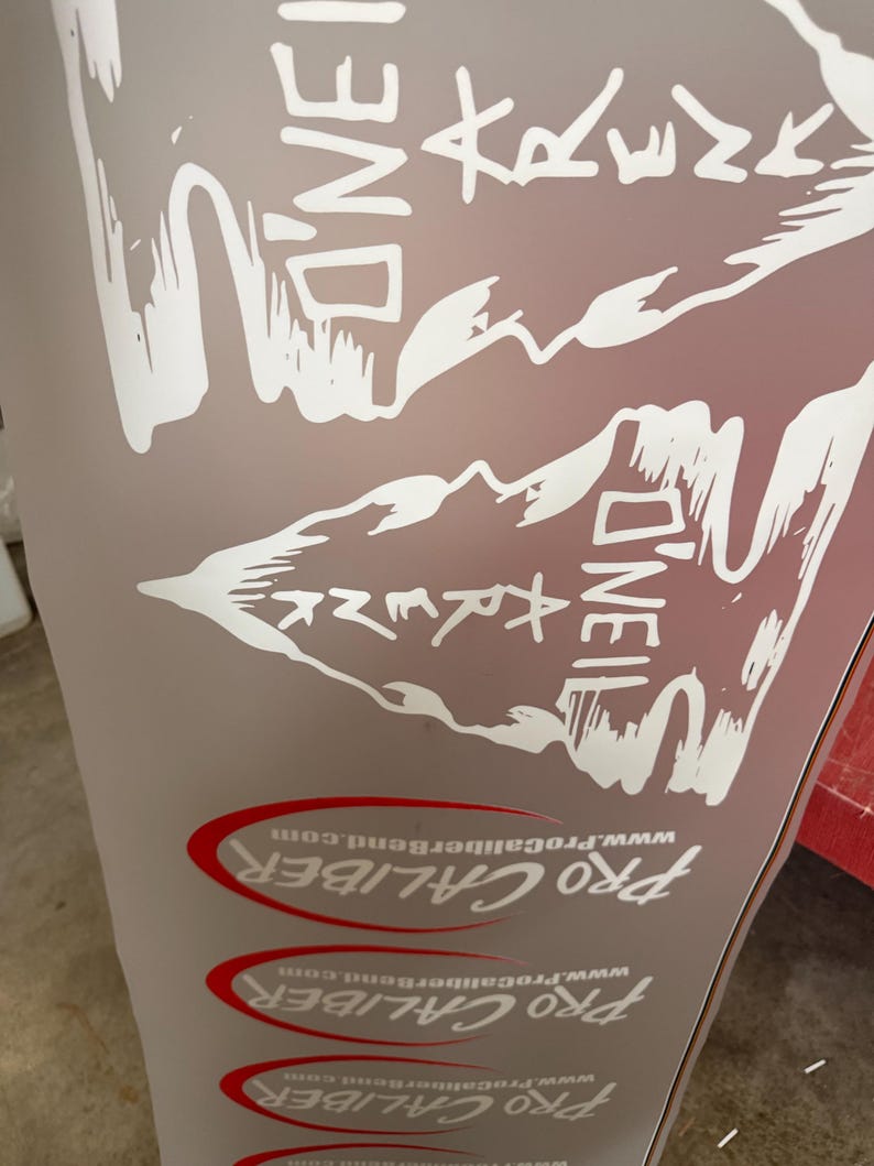 May include: A light brown surface featuring large white graphic text that reads O'NEIL KRENK in a stylized font. Below this, there are three stacked red oval outlines containing the white text PRO CALIBER and the website www.ProCaliberBend.com.