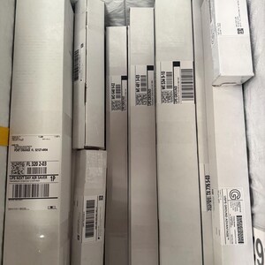 May include: A collection of rectangular white cardboard shipping boxes arranged vertically. Each box features various shipping labels with barcodes, QR codes, and text including UPS Ground, UPS Next Day Air Saver, and USPS Ground Advantage markings.