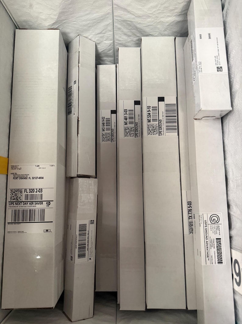 May include: A collection of rectangular white cardboard shipping boxes arranged vertically. Each box features various shipping labels with barcodes, QR codes, and text including UPS Ground, UPS Next Day Air Saver, and USPS Ground Advantage markings.