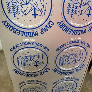 May include: A white sheet featuring multiple blue circular stickers. Each sticker displays a tent, pine trees, and a cross with the text "CAMP MIDDLEBURY", "FAITH IN THE WILDERNESS", "GOOD HOPE BAPTIST CHURCH", "WHERE FAITH TAKES ROOT", and "VBS 2026".