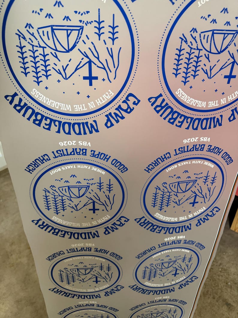 May include: A white sheet featuring multiple blue circular stickers. Each sticker displays a tent, pine trees, and a cross with the text "CAMP MIDDLEBURY", "FAITH IN THE WILDERNESS", "GOOD HOPE BAPTIST CHURCH", "WHERE FAITH TAKES ROOT", and "VBS 2026".