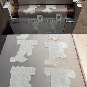 May include: A white industrial conveyor belt machine processing a transparent film sheet printed with repeating white graphic designs of banana peels and the text "BYKBY 33" and "MIFOEK2 38" in a grid layout on a flat surface.