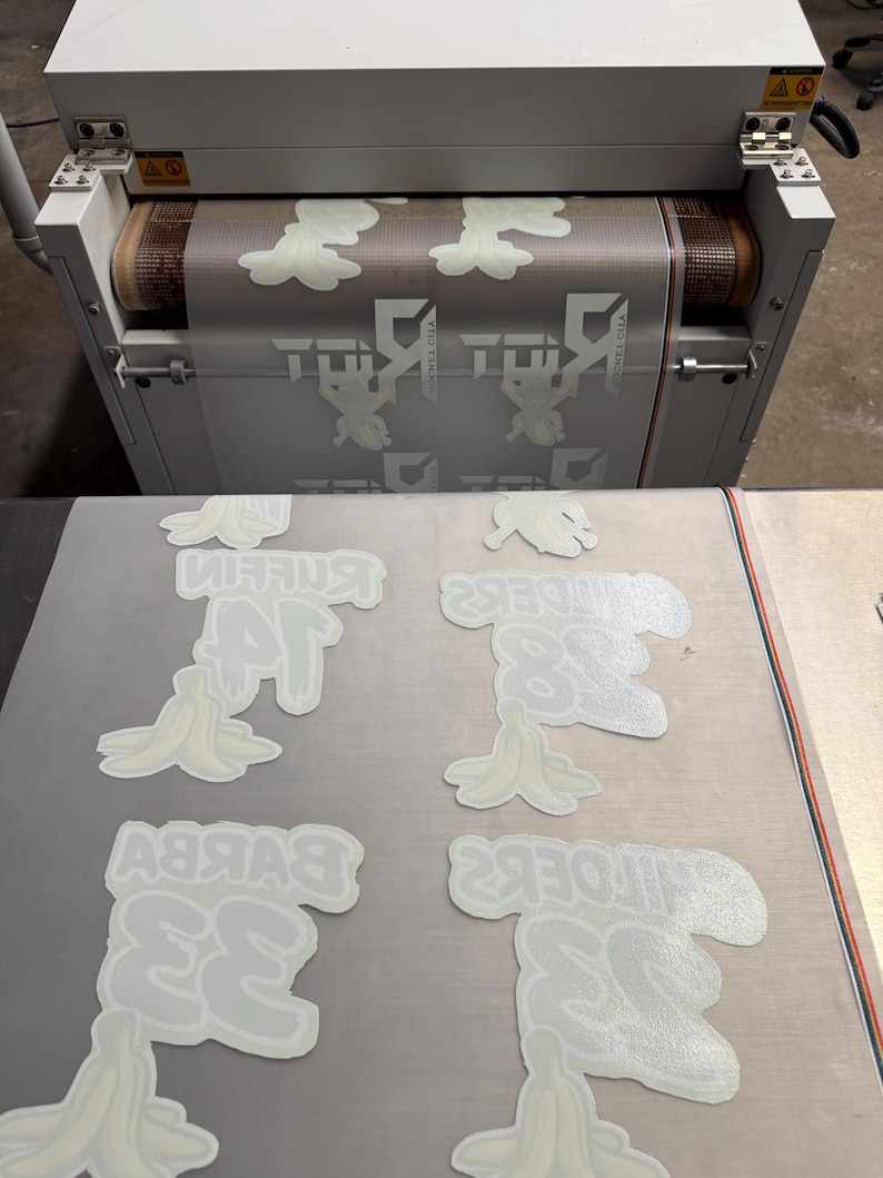 May include: A white industrial conveyor belt machine processing a transparent film sheet printed with repeating white graphic designs of banana peels and the text "BYKBY 33" and "MIFOEK2 38" in a grid layout on a flat surface.