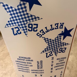 May include: A white poster featuring large blue stars and blue gingham patterned stars. The text includes "RETTES", "26 27", and a list of titles such as "CAPTAIN", "PRESIDENT", "ASST. DIRECTOR", "DIRECTOR", "SECRETARY", "TREASURER", and "LIEUTENANT".