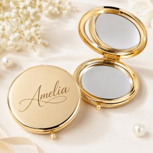 Pu&ograve; includere: Due specchietti da borsa rotondi in metallo dorato. Uno specchio &egrave; chiuso e mostra il nome Amelia in un'elegante scrittura corsiva sul coperchio. L'altro specchio &egrave; aperto, rivelando due superfici riflettenti all'interno. Finitura lucida.