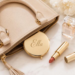 Pu&ograve; includere: Una borsa beige testurizzata con uno specchietto compatto in metallo dorato inciso con il nome Ella, un piccolo flacone di profumo in vetro con tappo trasparente e un rossetto dorato con la tonalit&agrave; color pesca esposta.