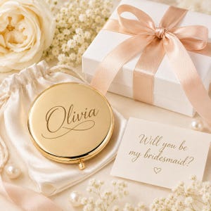 Pu&ograve; includere: Uno specchietto da borsa rotondo color oro inciso con il nome Olivia in corsivo. Lo specchio &egrave; appoggiato su un sacchetto di raso bianco con coulisse accanto a una scatola regalo bianca legata con un nastro color pesca e un biglietto con la scritta Will you be my bridesmaid?