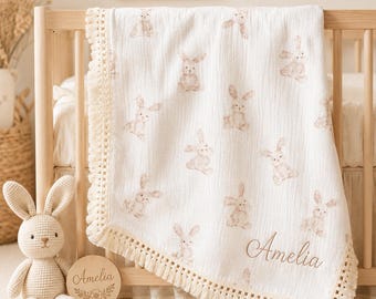 Può includere: Una coperta per neonati in mussola bianca con un motivo ripetuto di coniglietti color marrone chiaro ad acquerello e un bordo con frange color crema. Il nome Amelia è ricamato in corsivo marrone nell'angolo in basso a destra.