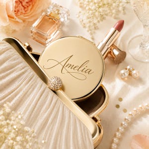 Pu&ograve; includere: Uno specchietto da borsa in metallo dorato con inciso il nome Amelia in corsivo, appoggiato all'interno di una pochette plissettata color crema. Lo specchietto &egrave; rotondo e presenta una piccola chiusura sferica testurizzata sul telaio.