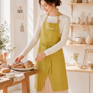 Könnte beinhalten: Eine chartreusefarbene Leinen-Schürze mit einer Vordertasche und dem in weißer Schreibschrift gestickten Namen Olivia auf der Brust. Die Schürze hat verstellbare Nackenbänder und ein Taillenband und wird über einem weißen Langarmshirt getragen.