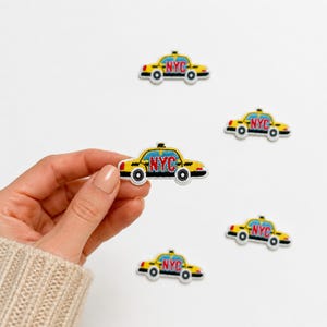 May include: Five embroidered patches shaped like yellow New York City taxi cabs. Each patch features a bright yellow body, blue windows, black wheels, and the letters NYC stitched in red across the side of the vehicle. The patches are small.