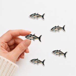 May include: Five small embroidered fish patches featuring a beige body, black outlines, and blue and yellow accents. Each patch depicts a fish in a swimming position with a pointed tail and fins, rendered in detailed thread work on a white background.