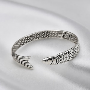 May include: A silver-toned open cuff bracelet shaped like a fish, featuring detailed scale textures along the band, a defined fish head at one end, and a tail fin at the other. The interior of the band displays a stamped 925 hallmark.