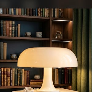 May include: A glossy off-white mushroom-shaped table lamp with a wide, rounded dome shade and a smooth, flared pedestal base. The lamp emits a soft, warm glow, illuminating the surrounding space while resting on a flat surface.