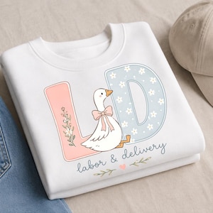 Könnte beinhalten: Ein weißes Sweatshirt mit Rundhalsausschnitt und einer Grafik einer weißen Gans mit rosa Schleife zwischen den Buchstaben L und D. Der Buchstabe L ist rosa mit Blumenakzenten, der Buchstabe D ist hellblau mit weißen Blumen. Text: labor & delivery.