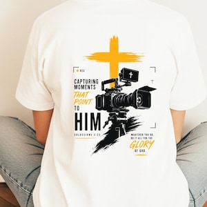 May include: A white t-shirt featuring a graphic of a black video camera on a tripod in front of a yellow cross. The text reads: CAPTURING MOMENTS THAT POINT TO HIM, COLOSSIANS 3:23, WHATEVER YOU DO, DO IT ALL FOR THE GLORY OF GOD.