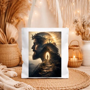May include: A white tote bag featuring a printed graphic of a bearded figure wearing a crown of thorns, with a path leading toward a glowing cave entrance and three crosses on a hill against a golden sunset sky, with two sturdy shoulder straps.