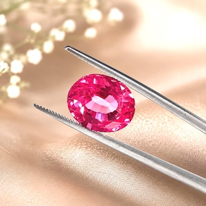 May include: A vibrant, oval-cut ruby gemstone with a deep red hue and multifaceted surface, held securely by a pair of silver-toned metal tweezers. The gemstone displays internal light reflections and a polished, transparent appearance.