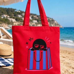 Könnte beinhalten: Eine leuchtend rote Tragetasche mit einer Grafik einer schwarzen, mürrisch aussehenden Figur, die aus einer blau-rot gestreiften Einkaufstasche hervorschaut. Das Design enthält kleine schwarze und rosa abstrakte Symbole um die Figur.